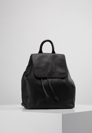 Sac à dos en cuir noir avec fermeture à rabat et cordon, posé sur une surface blanche devant un fond uni gris clair.