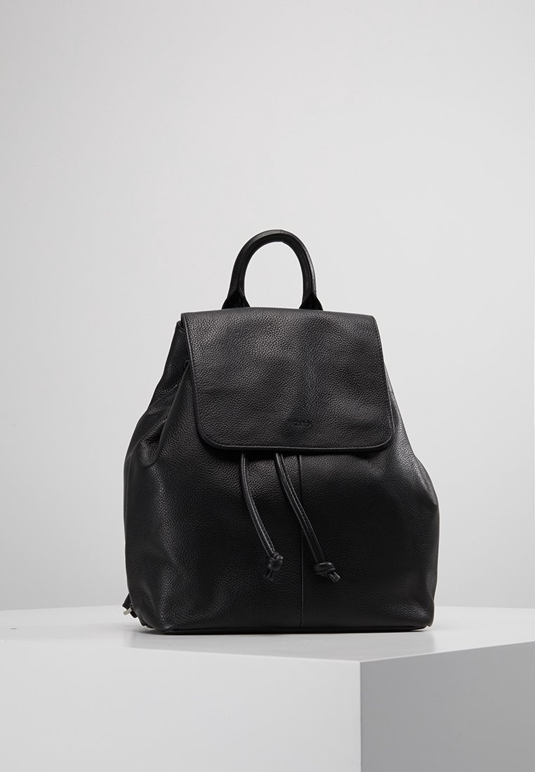 Sac à dos en cuir noir avec fermeture à rabat et cordon, posé sur une surface blanche devant un fond uni gris clair.