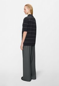 Mladá osoba s krátkymi blond vlasmi oblečená v čiernej krátkorukávovej polo košeli s tenkými horizontálnymi pruhmi a širokými sivými nohavicami, stojaca bokom.