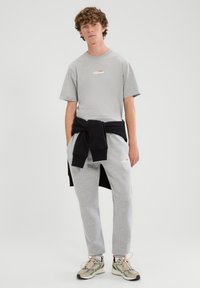 Grijs katoenen T-shirt met logo, gecombineerd met grijze joggers en een zwarte trui die om de taille is gebonden. Gecombineerd met sneakers met patroon.