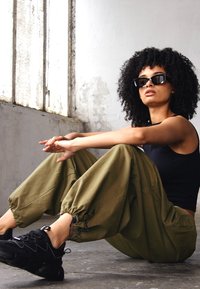 Top negro sin mangas recortado, pantalones cargo verde oliva con cordón y zapatos deportivos negros. El sujeto lleva puestas unas gafas de sol negras de gran tamaño, sentado contra una pared.