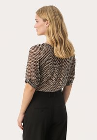 Genomskinlig topp med zigzag-mönster i brun, crème och svart; avslappnad passform med uppvikt ärm. Kombinerad med högmidjade svarta byxor.