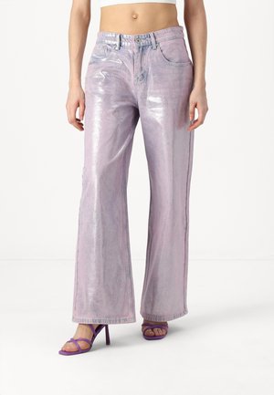 Vrouw draagt glanzende lichtpaarse wijde jeans en paarse sandalen met hak en bandjes, staand tegen een effen witte achtergrond.