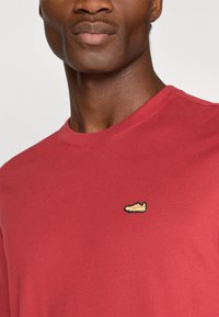 Κόκκινη βαμβακερή μπλούζα με στρογγυλή λαιμόκοψη, με ένα μικρό κομμένο λογότυπο από sneaker σε κίτρινο και μαύρο στην αριστερή πλευρά του στήθους.