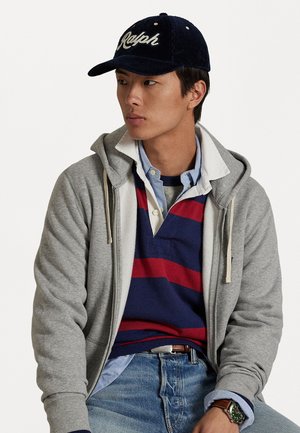 Jovem vestindo um boné azul-marinho "Ralph", um hoodie cinza com fecho, uma camisa de rugby listrada em azul-marinho e vermelho, uma camisa de colarinho azul-claro, jeans azuis e um relógio verde.