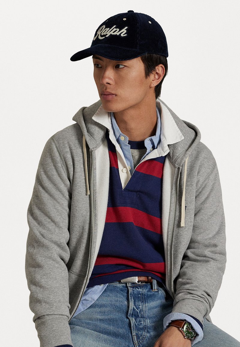 Ung mann iført en marineblå "Ralph" caps, grå hettegenser med glidelås, stripet marineblå og rød rugbyshirt, lyseblå skjorte med krage, blå jeans og et grønt armbåndsur.