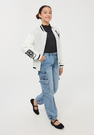 Giacca varsity grigia e bianca con dettagli neri, dolcevita nero, jeans cargo blu chiaro con tasche e ballerine nere.