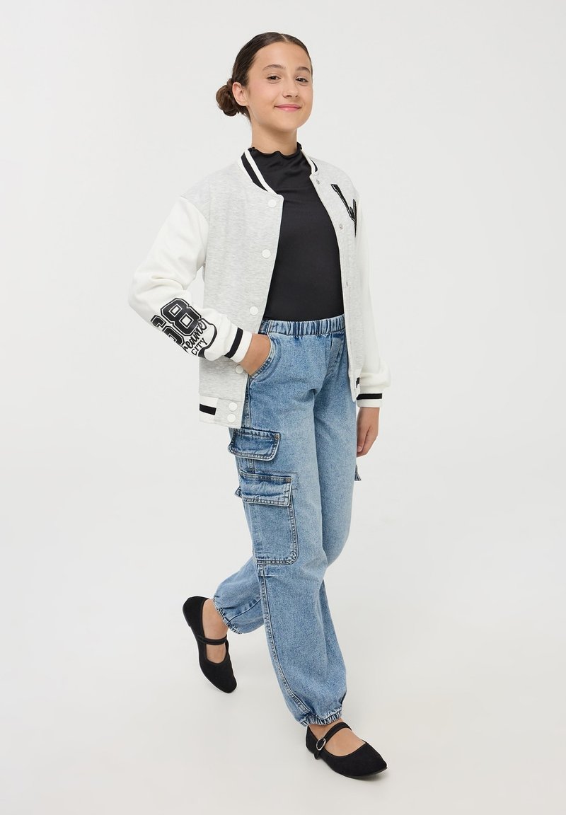 Giacca varsity grigia e bianca con dettagli neri, dolcevita nero, jeans cargo blu chiaro con tasche e ballerine nere.