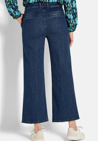 Jeans à jambe large en denim foncé avec une finition propre, dotés de poches arrière et d'une taille mi-haute, associés à des baskets de couleur claire.