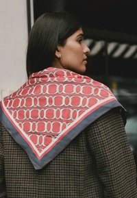 Sciarpa di seta rossa e bianca con motivo, di forma triangolare con bordo blu a contrasto, indossata sopra un cappotto a scacchi.