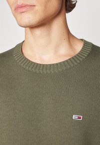 Pull en tricot vert olive avec un col rond côtelé et un écusson logo brodé en rouge, blanc et bleu sur la zone de la poitrine gauche.