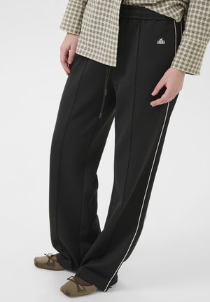 En person står i svarta träningbyxor med vita ränder, iklädd en rutigt topp och bruna skor, och visar upp en avslappnad outfit.