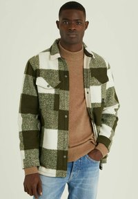 Groen en witte geblokte flanellen jas met twee voorkruiszakken, gedragen over een beige geribde trui, gecombineerd met lichtblauwe denim jeans.