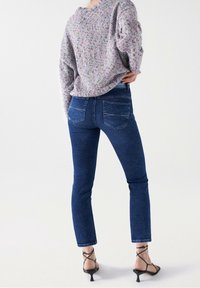 Pull en tricot gris tacheté ; jean bleu ajusté avec poches arrière ; escarpins noirs à lanières ; personne de dos, montrant les détails de la tenue.