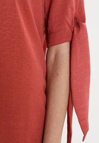 Halenka barvy rezavé s texturovanou látkou, krátkými rukávy a zavazovacími detaily na manžetách pro nastavitelný střih a zdůraznění.