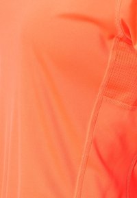 Ljus orange atletisk tröja med en slät textur, som har andningsbara meshnätpaneler och sömmar längs kanterna.