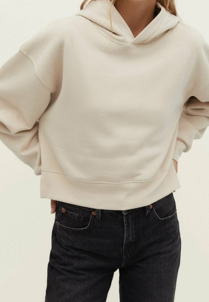 Sweat à capuche beige à manches longues porté par une personne les mains dans le dos, assorti à un jean en denim noir.