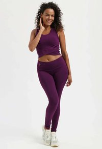 Lila sportliches Crop-Top mit passenden Leggings, die ein figurbetontes Design mit glatter Textur und dezenten Markenakzenten zeigen. Weiße Turnschuhe vervollständigen das Outfit.