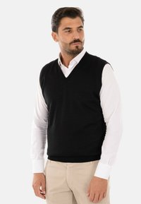 Gilet nero lavorato a maglia con scollo a V, indossato sopra una camicia bianca con bottoni. Il tessuto appare liscio con una silhouette aderente.