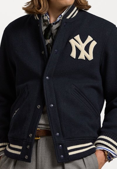 Marine uld varsity jakke med fremtrædende cremefarvet NY-logo, stribet krave og manchetter, trykknapper og sidelommer. Strukturdetalje i stoffet er synlig.