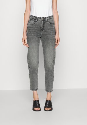 Person trägt hochtaillierte graue Jeans, weißes ärmelloses Oberteil und schwarze offene Slip-On-Schuhe und steht vor schlichtem Hintergrund.