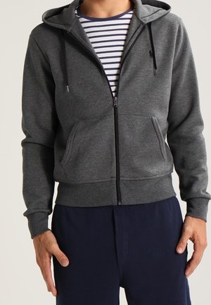 Man wearing a dark gray zip-up hoodie over a white and navy striped t-shirt with navy pants, standing with hands near pockets.