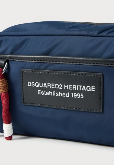 Τσάντα DSQUARED2 HERITAGE σε ναυτικό μπλε με μαύρη ετικέτα που αναγράφει "Established 1995" και κόκκινη και λευκή θηλιά φερμουάρ σε σχοινί.