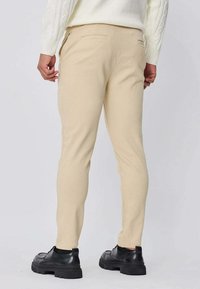 Pantalon beige sur mesure, coupe slim, avec une texture lisse, poches arrière, accompagné de chaussures en dentelle noires.