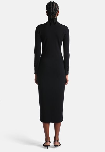 Femme aux cheveux tressés portant une robe midi noire moulante à manches longues et des sandales noires à talons bas, debout de dos sur un fond blanc.
