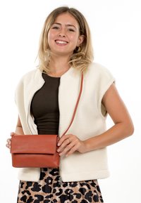 Jonge vrouw die lacht, gekleed in een witte mouwloze jas, een donker shirt, een rokje met luipaardprint en een kleine bruine leren crossbodytas vasthoudt.