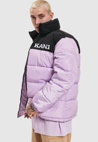 Puffjakke med lavendel farve og sort krave, med KANI-logo på brystet, lynlås lukning og ribbede manchetter.