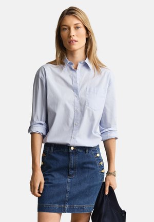 Femme portant une chemise bleu clair boutonnée avec les manches retroussées et une jupe en jean bleu avec des boutons dorés, tenant une veste foncée dans la main droite.