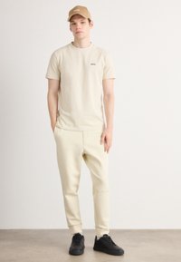 Beige Baumwoll-T-Shirt mit kleinem schwarzen "BOSS"-Logo, kombiniert mit hellbeigen Jogginghosen und schwarzen Turnschuhen; Träger befindet sich in einer neutralen Umgebung.
