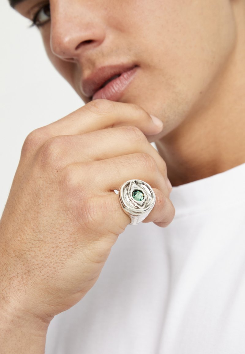 Anillo de plata con una banda texturizada y una piedra preciosa verde central, llevado en una mano sobre un fondo blanco liso.