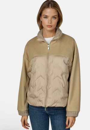 Jonge vrouw draagt een beige jas met rits, gebreide kraag en manchetten, gecombineerd met een blauwe spijkerbroek, tegen een effen witte achtergrond.