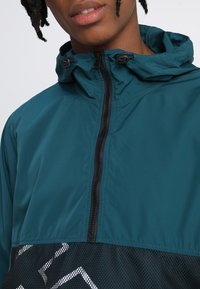 Tyrkysová větrovka s kapucí, s černým zipem a síťovanou spodní částí, která zobrazuje geometrický bílý vzor.