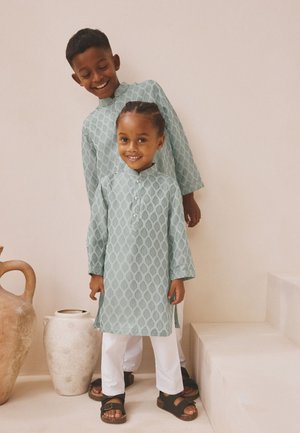 Deux enfants souriants portant des tuniques bleu clair à motifs assortis avec des pantalons blancs, debout à l'intérieur près de grands pots en argile et des escaliers.