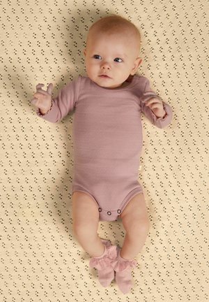 Bébé allongé sur une couverture beige à motifs, portant un body rose clair à manches longues et des chaussettes à volants assorties, regardant légèrement sur le côté.