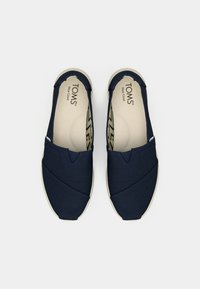 Laivaston väriset kangaslakkarit, joissa on ristikkäissuunnittelu päällä, beigepohja ja hienovarainen tekstuuri pohjassa, jossa on TOMS-logotypointi.