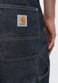 Ciemne ogrodniczki z denimu z kontrastowymi żółtymi szwami i kwadratową metką Carhartt. Posiadają kieszenie i trwały, teksturowany materiał.