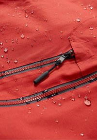 Vandafvisende rødt stof med dråber; har en sort lynlåslomme og tekstureret lynlåstræk. Glat overflade med mat finish.