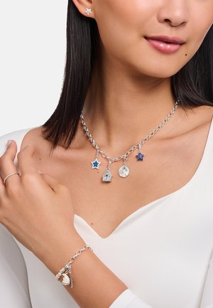 Silberne Kettenhalskette mit Stern- und Hausanhängern, die blaue Akzente aufweist. Das Armband hat Perlen und Charms und wird auf einer nackten Schulter getragen.