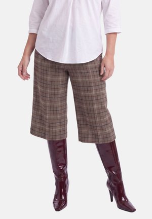 Vrouw die bruine geruite wijde cropped broek draagt, een wit overhemd en kniehoge donkerrode laarzen met hak.