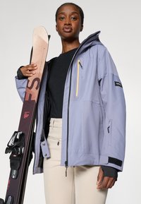 Hiihtotakki vaalean lilassa, jossa on keltainen vetoketjupikku, materiaalin tekstuuri, säädettävät hihansuut, yhdistettynä suksiin ja mustaan toppiin.