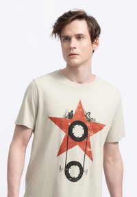 Lichtbeige t-shirt met een groot rood sterontwerp met zwarte grafische elementen in het midden. Korte mouwen, ronde halslijn, de stof lijkt zacht te zijn.