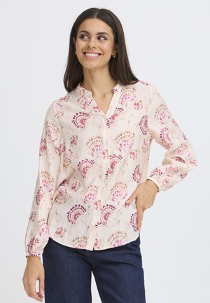 Junge Frau trägt eine hellrosa Blumenbluse mit Knöpfen und dunkle Jeans, lächelt und schaut zur Seite, die Hand in die Hüfte gestützt.