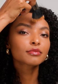 Femme aux cheveux bouclés appliquant un fond de teint sur le front avec un pinceau noir et orange, portant des boucles d'oreilles créoles en or.