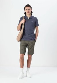 Kortærmet marineblå polo skjorte med hvid krave, parret med olivengrønne shorts. Modellen bærer en beige taske og har hvide sneakers og sokker på.