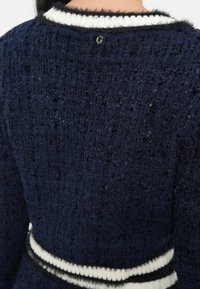 Primo piano della schiena di una persona che indossa un maglione testurizzato blu navy con bordo morbido bianco e nero al colletto e in vita, con una piccola spilla dorata con la lettera "G".