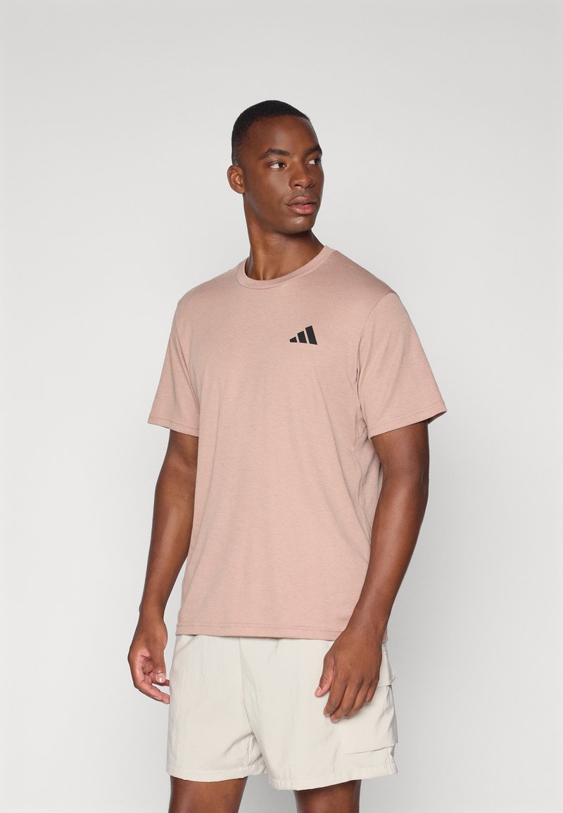 Lys rosa t-skjorte med korte ermer laget av myk stoff, med en svart logo på venstre bryst, kombinert med beige shorts.